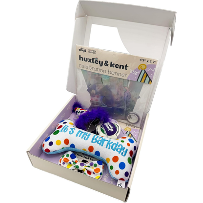Open box with a colorful polka dot dog toy, purple feather, and a celebration banner inside.